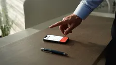 Ruka sahá po chytrém telefonu na stole, vedle leží pero, pracovní prostředí.