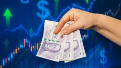 Ruka držící české bankovky před grafem s finančními symboly a šipkami vzhůru.