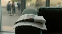 Noviny položené na opěrce pohovky v moderním interiéru s oknem na pozadí.