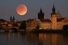 Pražský noční panoramat s úplňkem a historickými budovami na břehu Vltavy.